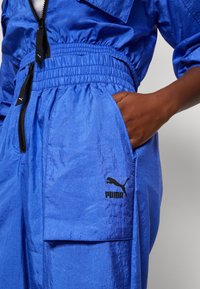 Pantalones de chándal azules con cinturilla elástica, que cuentan con un bolsillo lateral y un logo negro de Puma. El material parece ligero y ligeramente texturizado.