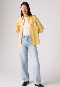 Camicia gialla a righe sopra una maglietta bianca attillata, abbinata a jeans blu chiaro larghi e scarpe nere. Outfit semplice e casual.