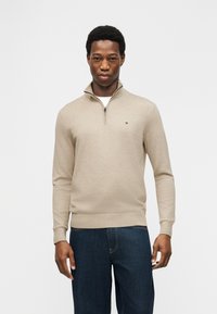 Νεαρός άνδρας με μπεζ πουλόβερ με φερμουάρ μέχρι το στήθος και σκούρο μπλε τζιν, στέκεται μπροστά σε απλό φωτεινό φόντο.