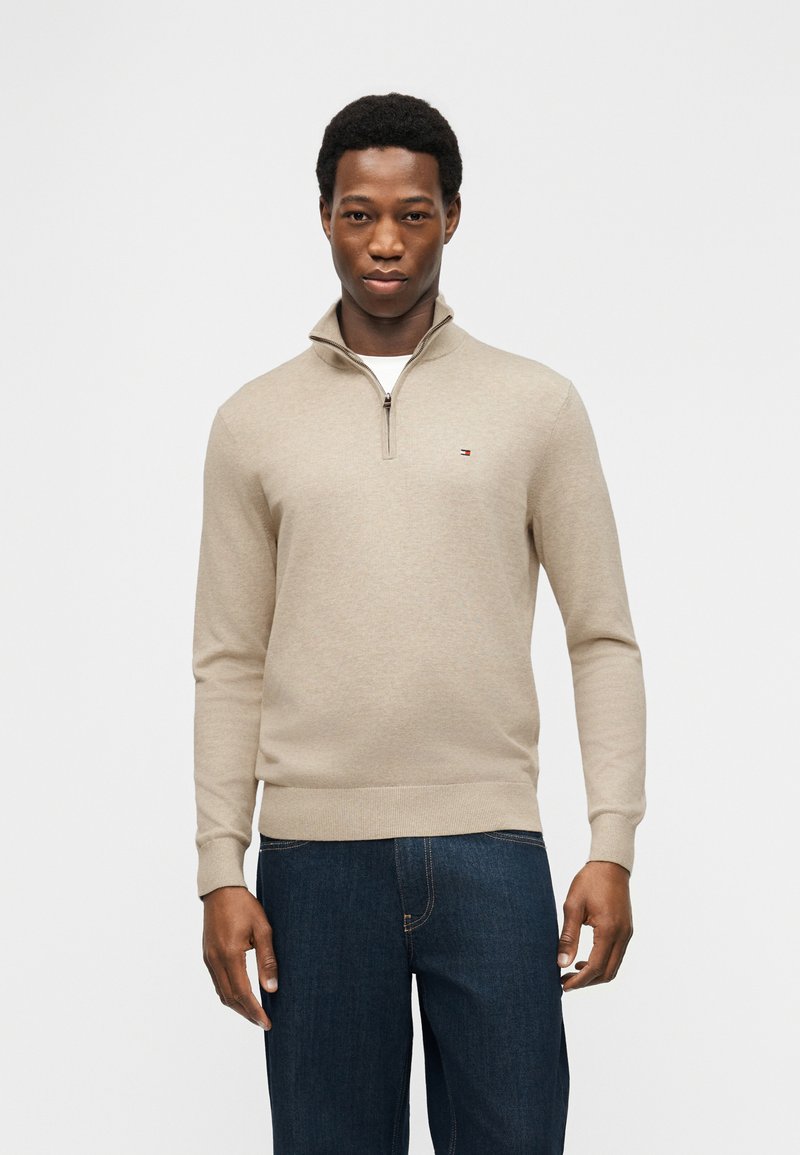 Νεαρός άνδρας με μπεζ πουλόβερ με φερμουάρ μέχρι το στήθος και σκούρο μπλε τζιν, στέκεται μπροστά σε απλό φωτεινό φόντο.