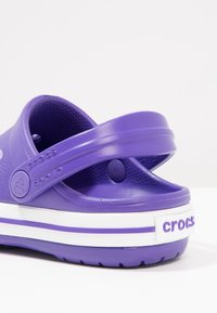 Crocs viola con suola strutturata, cinturino regolabile e striscia bianca sulla suola. Include il marchio sul tallone.