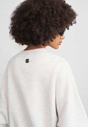 Gaiši pelēks sudraba krekls ar mīkstu tekstūru, pārāk liels piegriezums un noapaļots kakls. Aizmugurē ir neliels melns logotipa plāksteris.