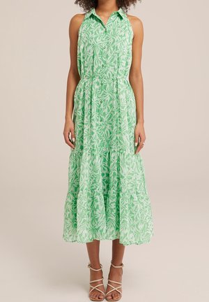 Robe midi verte sans manches avec motif feuilles blanches, boutonnée sur le devant, col et jupe à volants, portée avec des sandales beiges à brides.