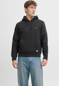 Felpa nera con cappuccio e logo tono su tono sul petto, tasca frontale a marsupio e polsini a costine. Indossata con jeans azzurri chiari.