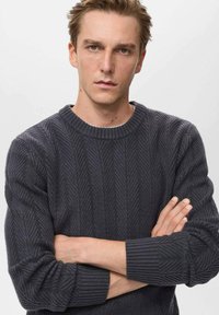 Suéter de punto gris oscuro con un patrón de espiga texturizado, cuello redondo y puños acanalados. El modelo tiene los brazos cruzados.