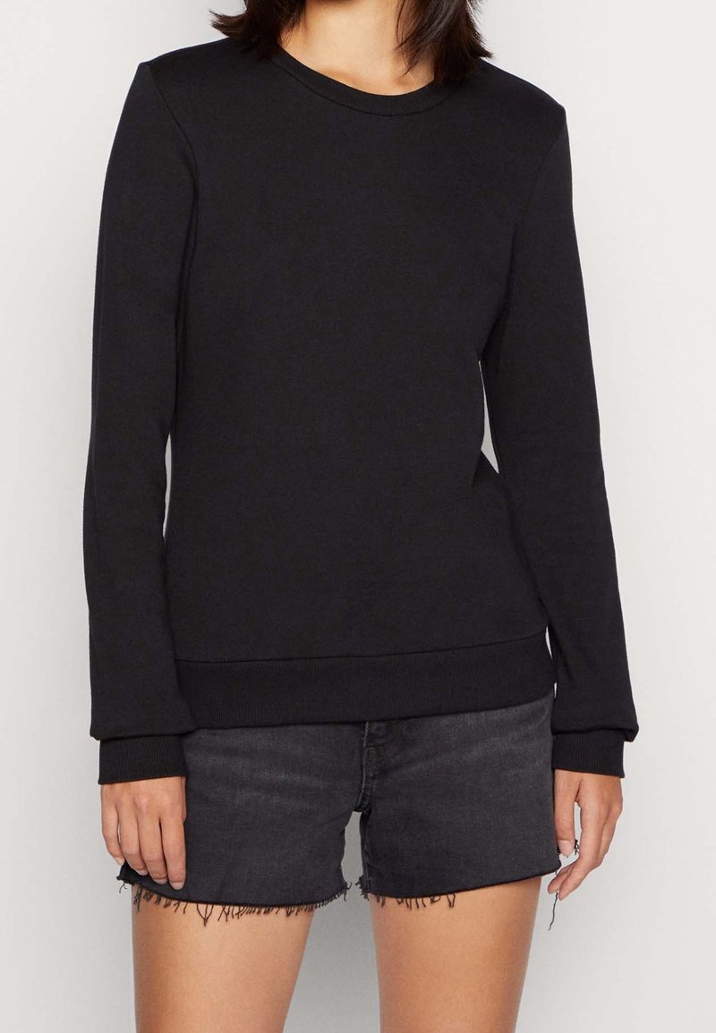 Femme portant un sweat-shirt noir uni à manches longues et un short en jean noir effiloché, debout devant un fond clair uni.