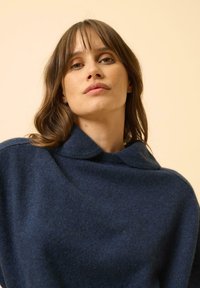 Námořnická modrá vlněná svetr s vysokým límcem a volným střihem. Má měkkou texturu a minimalistický design. Hladký povrch bez viditelných vzorů.