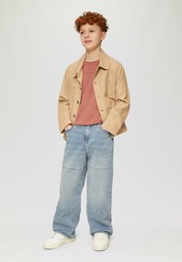 Tan gekleurde jas over een roestrode t-shirt, gecombineerd met lichtblauwe wijde jeans. Witte sneakers maken de outfit compleet.