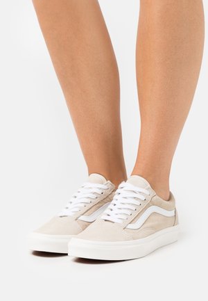 Zapatillas de ante beige con cordones blancos y una franja lateral blanca. Punta redondeada y suela de goma con detalles texturizados. Diseño de corte bajo.