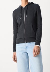 Schwarze Zip-Hoodie aus weichem Material, mit einem vorderen Reißverschluss, zwei Taschen und einer verstellbaren Kapuze. Getragen mit hellblauen Jeans.