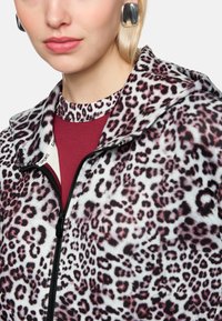 Leopardenmuster-Reißverschlussjacke aus weichem Stoff mit Kapuze. Mit pinken und burgunderroten Akzenten, unter einem Rundhalsausschnitt-Shirt getragen.