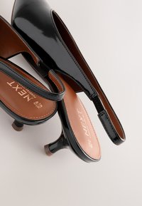 Mustad lakknahast sling-kingad madala kontsaga ja pruuni voodriga, millel on reljeefse logoga ning kontrastse äärega detailid.