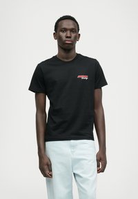 Μαύρη βαμβακερή μπλούζα με κοντά μανίκ�ια, στρογγυλή λαιμόκοψη, με ένα κόκκινο και λευκό γραφικό λογότυπο στην αριστερή πλευρά του στήθους. Φοριέται με ανοιχτό γαλάζιο παντελόνι.