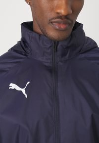 Marineblå windbreaker-jakke lavet af glat, letvægtsstof. Har en høj krave, fuld lynlås og et hvidt Puma-logo på venstre bryst.