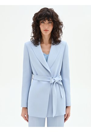 Donna con capelli ricci scuri che indossa un blazer azzurro chiaro con cintura e pantaloni abbinati, su uno sfondo bianco uniforme.