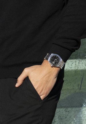 Montre-bracelet en argent avec un cadran noir circulaire, un bracelet en acier inoxydable et des marqueurs lumineux, posée sur une main reposant dans une poche noire.