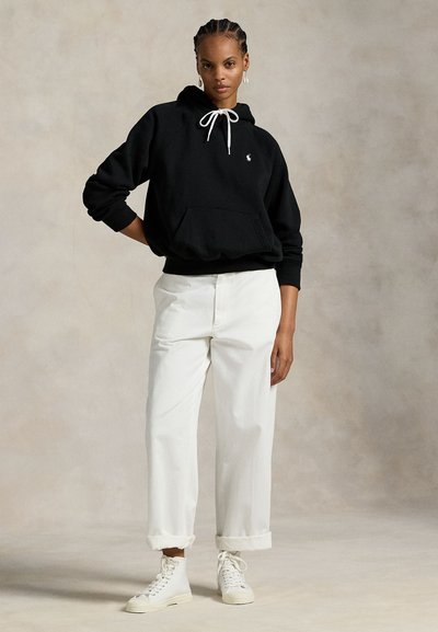 Vrouw staand, gekleed in een zwarte hoodie met witte trekkoorden, witte hoge taille broek met opgerolde pijpen en witte hoge sneakers.