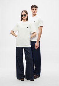 Άσπρα βαμβακερά t-shirt με μαύρο λογότυπο στο στήθος, συνδυασμένα με σκούρα τζιν παντελόνια με φαρδύ πόδι. Το μοντέλο φοράει γυαλιά ηλίου και στέκεται μπροστά.