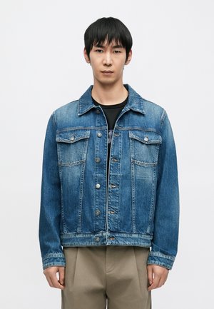 Blue denim jacket with a classic collar, button closure, two chest pockets, and a relaxed fit. Features a faded texture and sewn accents.