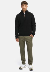 Zwarte gebreide trui met een hoge kraag en een halve rits; gecombineerd met olijfgroene broek en witte sneakers met groene accenten.