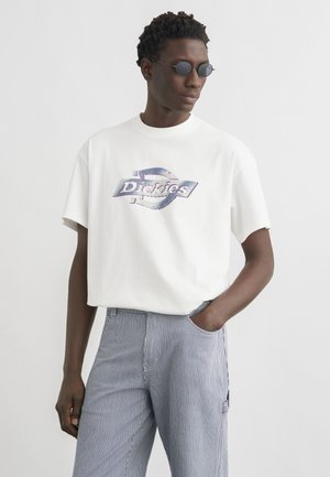 Vyras dėvi apvalius saulės akinius, baltą laisvo silueto marškinėlį su „Dickies“ logotipu ir mėlynas dryžuotas kelnes, viena ranka kišenėje.