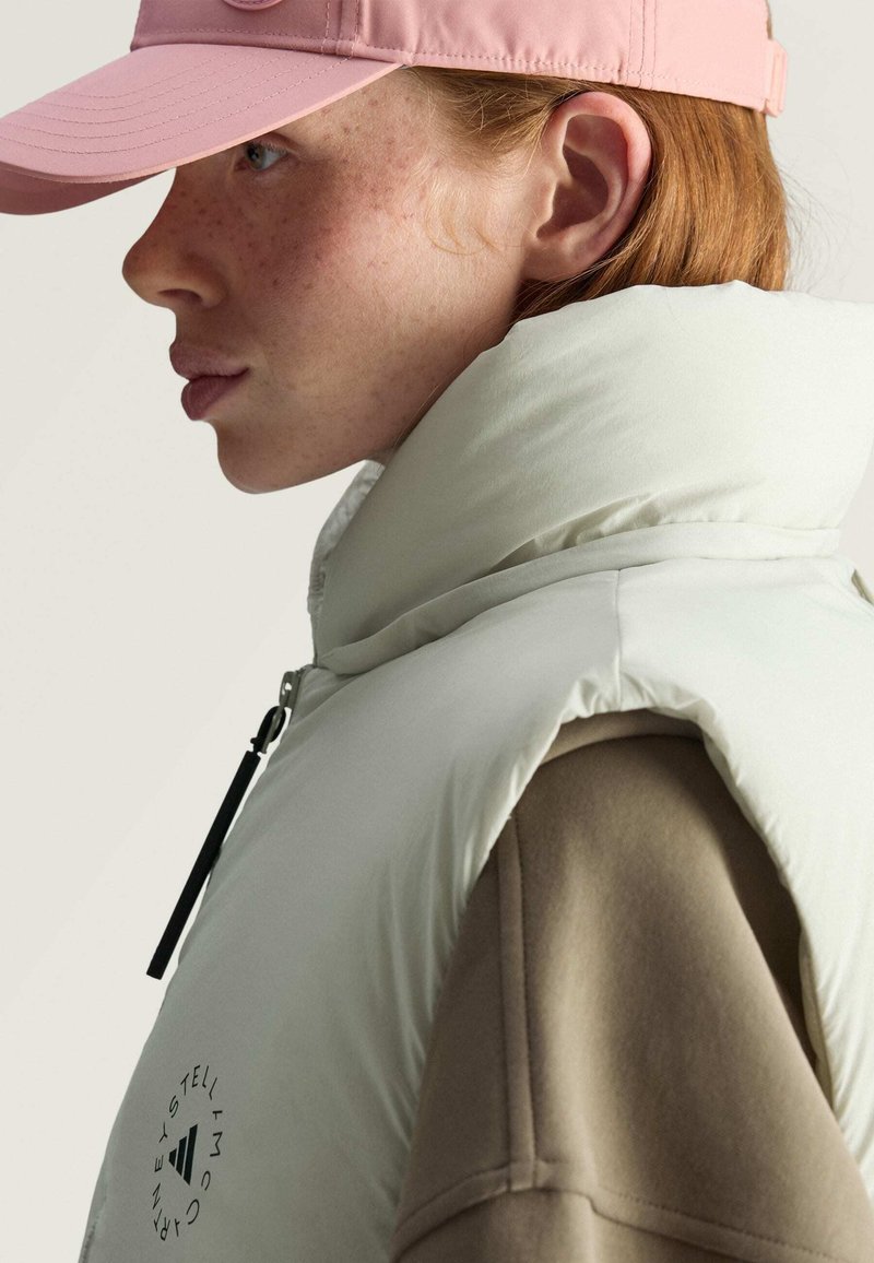 Chaleco acolchado verde claro con cuello alto, frente con cremallera y acentos negros. Lleva puesto sobre una blusa de manga larga beige, combinado con una gorra rosa.