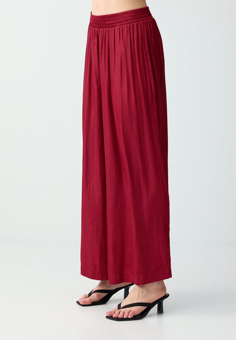Femme portant un pantalon rouge taille haute, ample et large, associé à des sandales noires à talons et fines lanières, sur un fond uni.