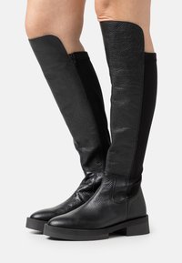 Cizme negre până la genunchi, cu un superior din piele texturată și un panou din material elastic. Talpă groasă din cauciuc și fermoar pe lateral pentru o purtare ușoară.