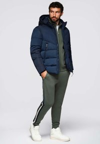 Giacca a vento blu navy con cappuccio, tuta verde slim-fit e scarpe da ginnastica bianche. La giacca presenta cuciture orizzontali e tasche laterali.