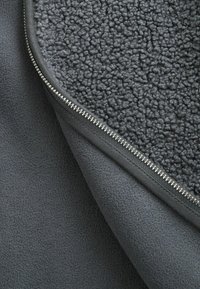 Coperta grigia in pile con una superficie interna testurizzata e soffice e uno strato esterno liscio, dotata di una cerniera argentata lungo il bordo.