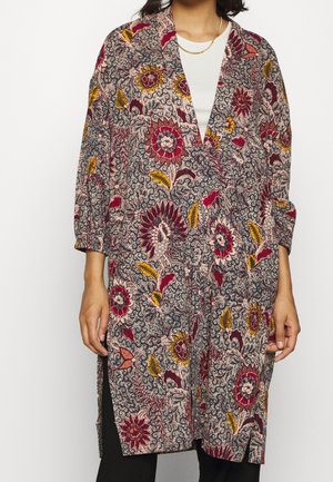 Woman wearing a long floral-patterned coat with red, yellow, and black flowers over a white top and black pants.