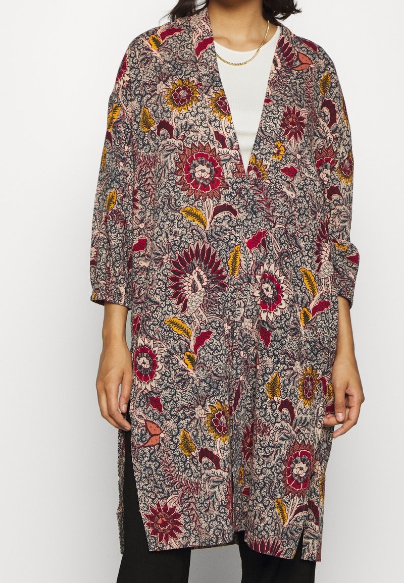 Kobieta ubrana w długi płaszcz w kwiatowy wzór z czerwonymi, żółtymi i czarnymi kwiatami, nosząca białą bluzkę i czarne spodnie.