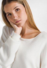 Weißer, strukturierter Pullover mit weitem Rundhalsausschnitt und gerippten Bündchen. Modell stützt das Kinn auf die Hand und zeigt Material- und Passformdetails.
