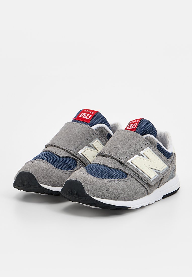 Baskets New Balance grises et bleu marine avec une sangle velcro, dotées d'une tige en maille, d'une semelle rembourrée et d'un logo 'N' blanc sur le côté.