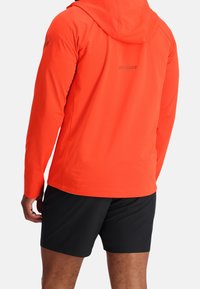 Homme portant une veste à capuche orange vif et un short noir, vu de dos, debout devant un fond blanc.