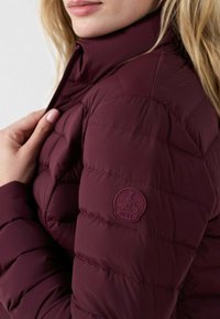 Bordeaux pufferjack met gestikt ontwerp, hoge kraag en zijrits. Heeft een rond logo-oproep op de mouw en een gladde textuur.