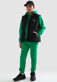 Černá zateplená vesta s dvěma kapsami přes zelenou mikinu a odpovídající kalhoty. Černé tenisky s šedými akcenty dotvářejí celý outfit.