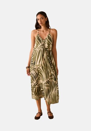 Vrouw draagt een mouwloze midi-jurk met groen- en beige tropisch bladmotief, met dunne bandjes, bruine platte schoenen en armbanden aan één pols.