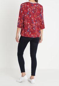 Blusa rossa floreale con maniche a tre quarti, caratterizzata da un motivo floreale multicolore. Abbinata a jeans skinny scuri e sneaker bianche.