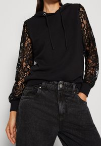 Felpa nera con maniche lunghe in pizzo, dotata di cappuccio regolabile e polsini a coste, abbinata a jeans in denim scuro.