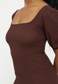 Femme portant une robe marron ajustée et texturée avec des manches courtes bouffantes et un décolleté carré, vue des épaules aux hanches.