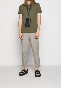 T-shirt verde oliva a maniche corte con un piccolo logo rosso, abbinato a pantaloni grigi chiari e larghi e sandali neri. Una borsa a tracolla nera pende da una tracolla.
