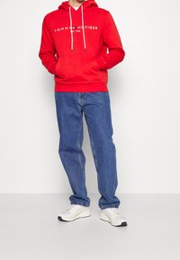 Sudadera de algodón roja con cordones blancos y bolsillo frontal, con el logo "TOMMY HILFIGER EST. 1985". Combinada con jeans de mezclilla azules y zapatillas blancas.