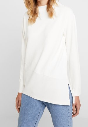 Femme portant un pull asymétrique blanc à manches longues avec des fentes latérales, associé à un jean bleu en denim sur un fond uni.