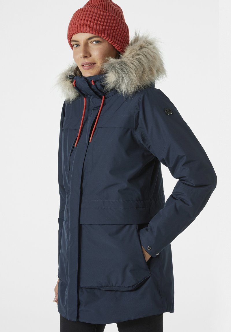 Navy waterproof parka with fur-trimmed hood, toggle adjusters, large front pockets, and snap cuffs, worn with a red knitted beanie.