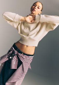 Žena v svetlom svetri a pruhovanej košeli viazanej okolo pása pózuje s rukami pri tvári, na dlani má napísané slovo "ÚSMEV", neutrálny pozadí.