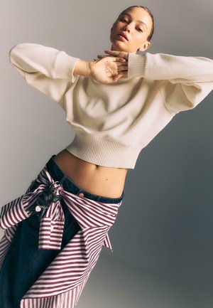 Kvinde med lys sweater og stribet skjorte bundet om livet poserer med hænderne tæt på ansigtet, ordet "SMILE" skrevet på håndfladen, neutral baggrund.