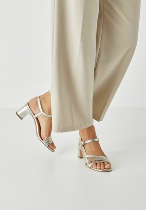 Sandales à talons argentés avec plusieurs brides, design à bout ouvert et talon carré. Portées avec un pantalon large beige clair.