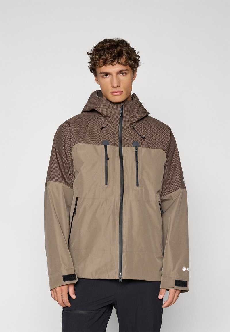 Giacca impermeabile marrone con parte superiore testurizzata, chiusura frontale con zip, due tasche sul petto con zip e cappuccio regolabile.
