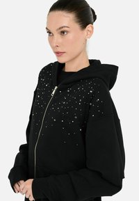 Modèle avec les cheveux coiffés en arrière portant un hoodie noir zippé orné d'embellissements scintillants, posant devant un fond blanc uni.